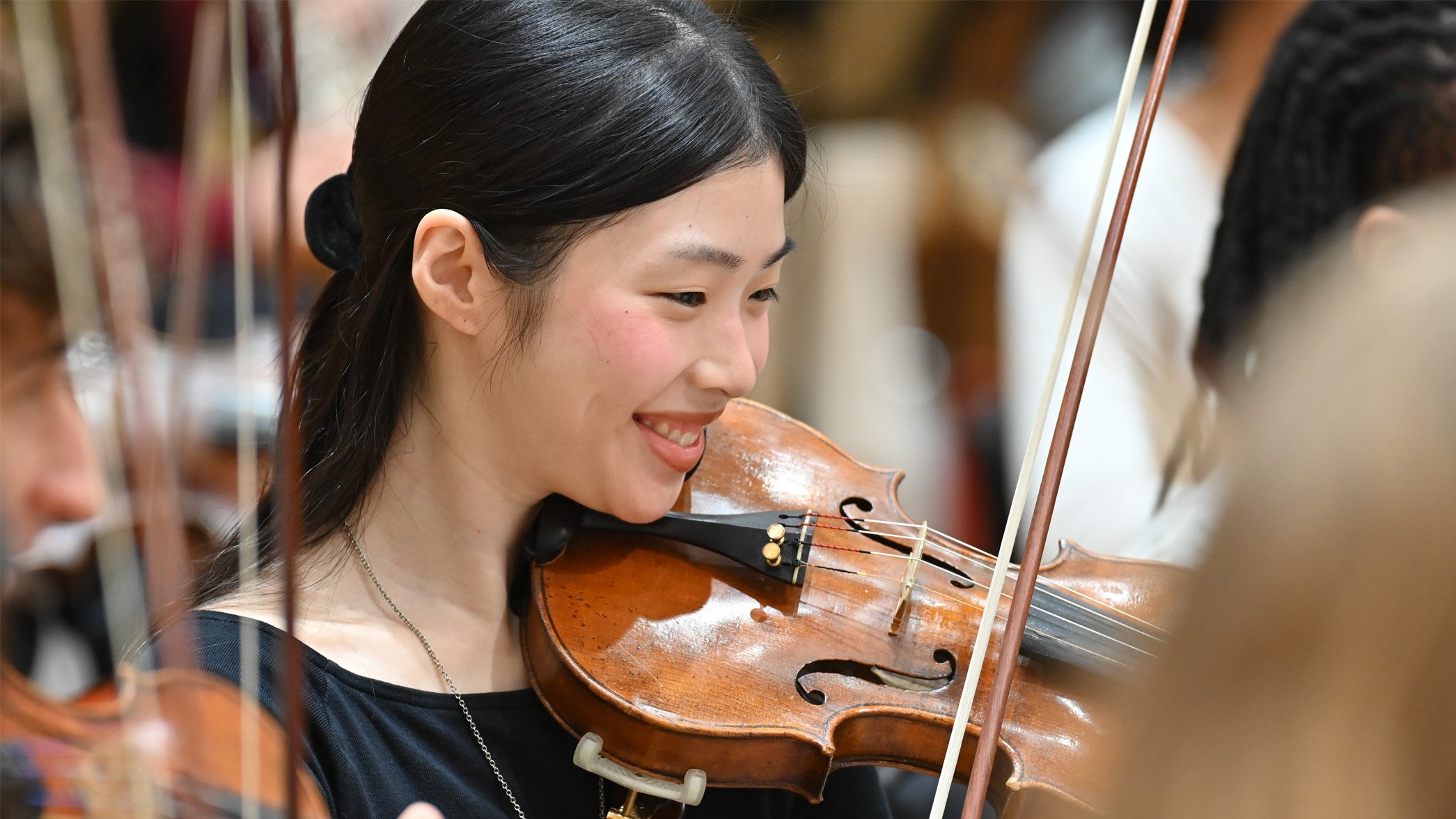 A female student playing the violin, smiling while playing in an orchestra rehearsal.